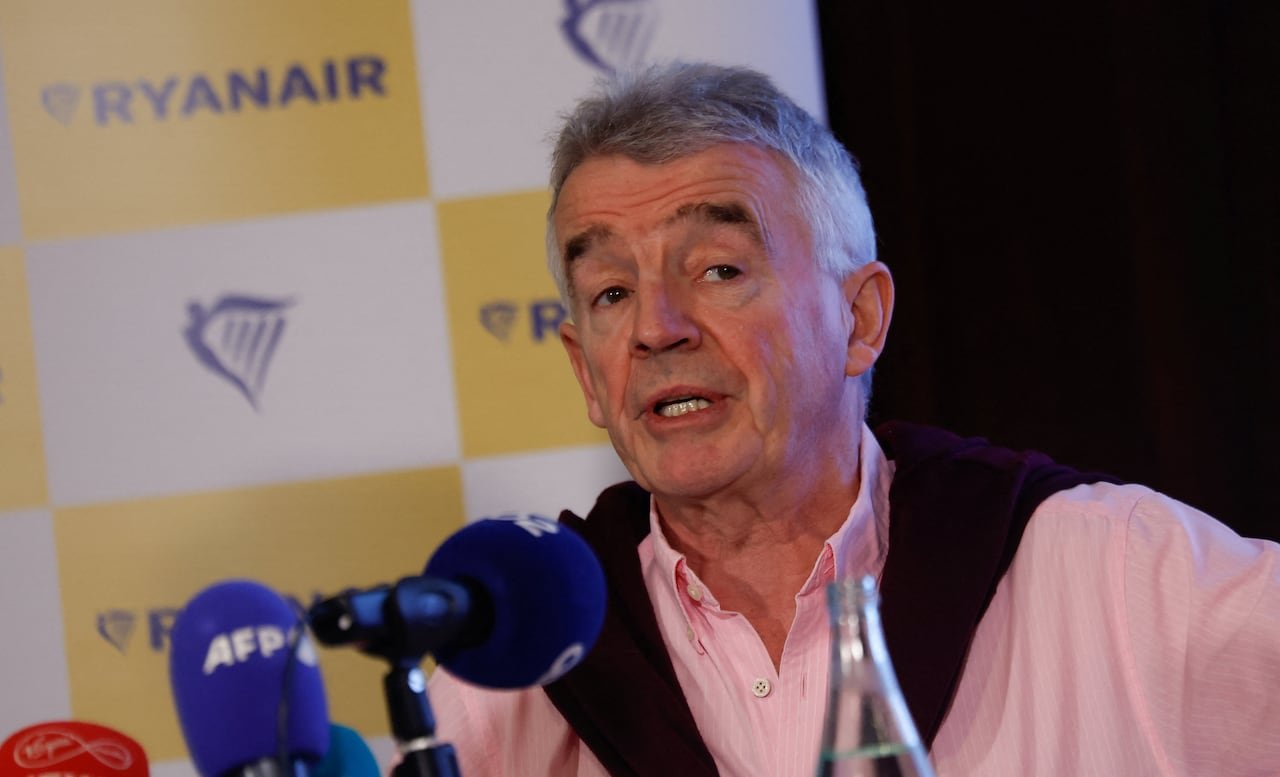 default-280 A man with white hair wearing a pale pink shirt talks in front of several microphones.
