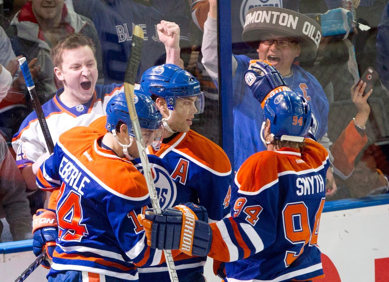 Three hockey players in orange-and-blue jerseys hug and celebrate while fans on the other side of the arena glass cheer wildly.