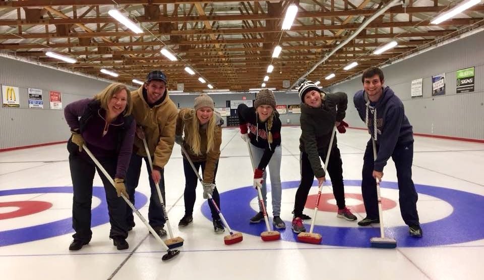 default-89 Two men and four women with sweeping brooms stand on the painted circles of a curling rink.