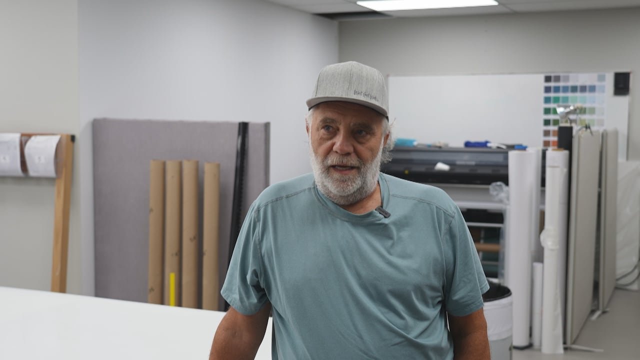 A man with a beard and a baseball cap surrounded by wallpaper rolls.