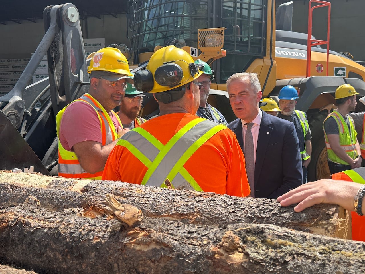 A man in a suit addressing mill workers surrounded by logs.